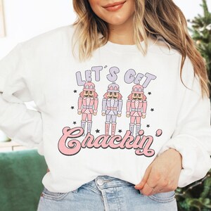 May include: White sweatshirt with the text "LET'S GET Crackin" in pink and purple. The graphic features three nutcracker figures in pink, purple, and blue uniforms. The sweatshirt is a long-sleeved crew neck.