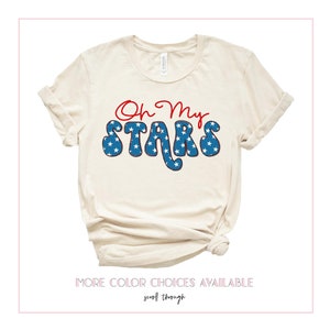 May include: A beige t-shirt with the text "Oh My Stars" in red and blue. The text is in a retro font and features white stars on a blue background.