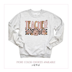 Puede incluir: Una sudadera gris con la palabra "TEACHER" impresa en letras rosas y marrones. El dise&ntilde;o incluye un patr&oacute;n a cuadros, un autob&uacute;s escolar, una mochila y un libro.