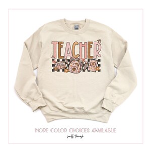 Puede incluir: Una sudadera beige con la palabra "TEACHER" impresa en letras rosas y marrones. El dise&ntilde;o incluye un autob&uacute;s escolar, una mochila y otros &uacute;tiles escolares con detalles florales.