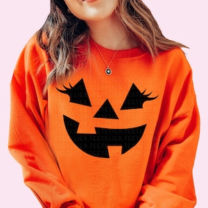 Może przedstawiać: Pomarańczowa bluza z nadrukiem czarnej twarzy jack-o'-lantern. Twarz ma rzęsy i szeroki uśmiech.