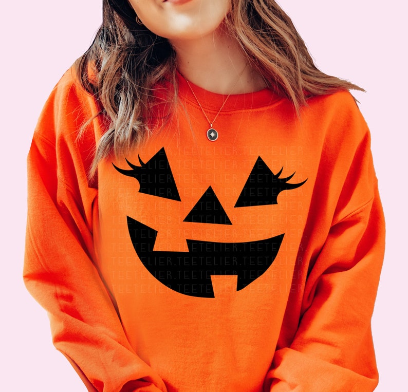 Może przedstawiać: Pomarańczowa bluza z nadrukiem czarnej twarzy jack-o'-lantern. Twarz ma rzęsy i szeroki uśmiech.