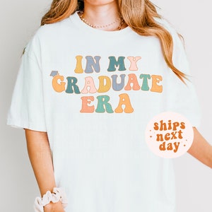 K&ouml;nnte beinhalten: Wei&szlig;es T-Shirt mit dem Text "In My Graduate Era" in bunter Retro-Schrift. Der Text ist in einem Regenbogen aus Farben, darunter Gelb, Orange, Rosa, Blau und Gr&uuml;n. Das Shirt wird von einer Person mit langen blonden Haaren getragen. Ein kleiner wei&szlig;er Kreis mit dem Text "ships next day" befindet sich in der unteren rechten Ecke des Bildes.