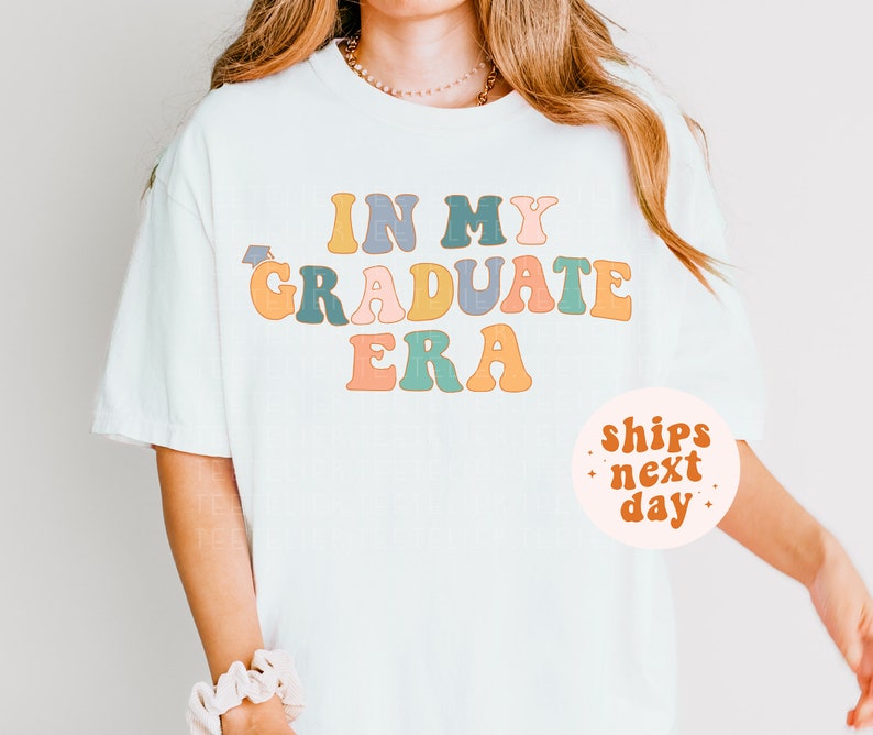 K&ouml;nnte beinhalten: Wei&szlig;es T-Shirt mit dem Text "In My Graduate Era" in bunter Retro-Schrift. Der Text ist in einem Regenbogen aus Farben, darunter Gelb, Orange, Rosa, Blau und Gr&uuml;n. Das Shirt wird von einer Person mit langen blonden Haaren getragen. Ein kleiner wei&szlig;er Kreis mit dem Text "ships next day" befindet sich in der unteren rechten Ecke des Bildes.