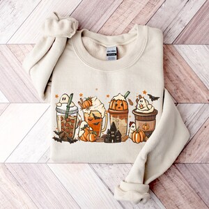 May include: A beige sweatshirt with a graphic print of four fall-themed coffee drinks with ghosts, bats, pumpkins, and stars. The text "Gildan" is visible on the tag.