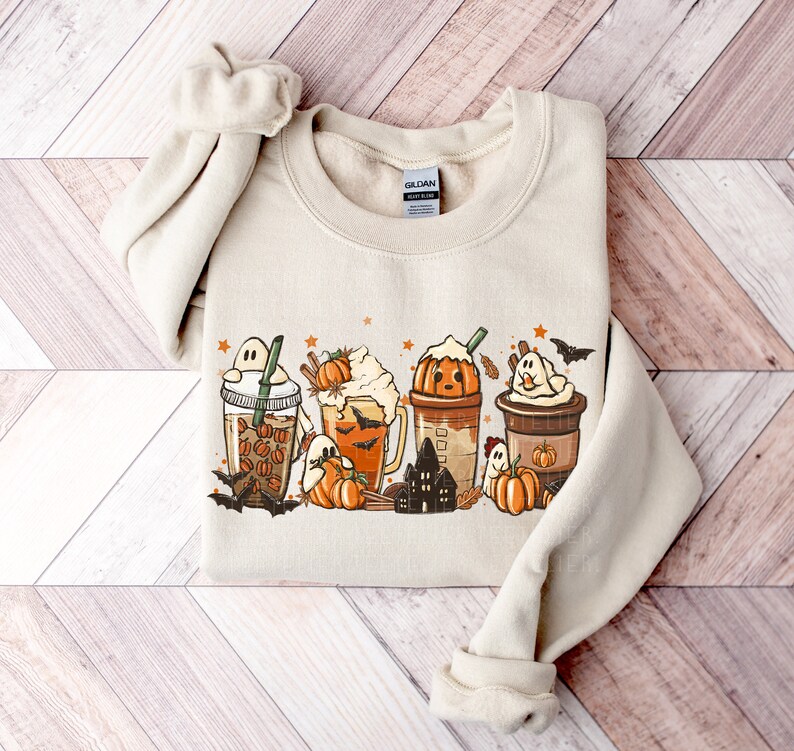 May include: A beige sweatshirt with a graphic print of four fall-themed coffee drinks with ghosts, bats, pumpkins, and stars. The text "Gildan" is visible on the tag.