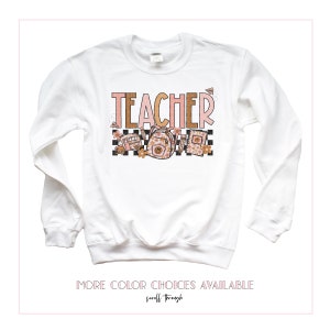 Puede incluir: Sudadera blanca de manga larga con la palabra "TEACHER" en letras rosas y marrones. El dise&ntilde;o incluye un patr&oacute;n a cuadros, un autob&uacute;s escolar, una mochila y l&aacute;pices. El texto "MORE COLOR CHOICES AVAILABLE" est&aacute; en la parte inferior de la imagen.