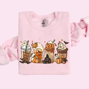 May include: A pink sweatshirt with a graphic of five fall-themed coffee drinks with ghosts, pumpkins, and bats. The drinks are topped with whipped cream, pumpkin spice, and sprinkles.