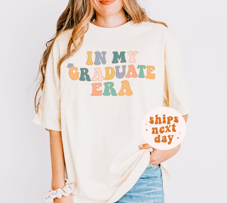 K&ouml;nnte beinhalten: Ein hellgelbes, &uuml;bergro&szlig;es T-Shirt mit dem Text "IN MY GRADUATE ERA" in bunten Retro-Buchstaben. Die Buchstaben sind in den Farben Rosa, Blau, Gr&uuml;n, Gelb und Orange gehalten.
