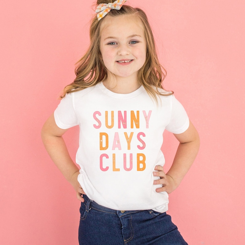 May include: A white t-shirt with a graphic print that says "SUNNY DAYS CLUB" in pink, orange, and yellow letters. The letters are stacked on top of each other and have a distressed look.