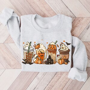 May include: A gray sweatshirt with a Halloween-themed design featuring four different fall-themed drinks with ghosts, pumpkins, and bats. The drinks are decorated with whipped cream, sprinkles, and cinnamon sticks.