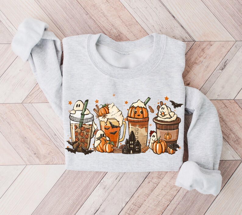 May include: A gray sweatshirt with a Halloween-themed design featuring four different fall-themed drinks with ghosts, pumpkins, and bats. The drinks are decorated with whipped cream, sprinkles, and cinnamon sticks.