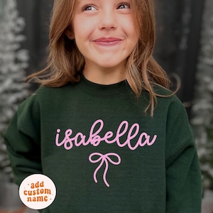 May include: A dark green sweatshirt with the name "isabella" in pink cursive lettering and a pink bow graphic. The sweatshirt has a round neck and long sleeves. A small, round sticker with the text "add custom name" is in the lower left corner.