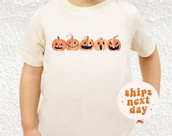 Könnte beinhalten: Cremefarbenes kurzärmeliges T-Shirt mit einer Reihe von fünf orangefarbenen Kürbissen mit unterschiedlichen geschnitzten Gesichtern. Die Kürbisse haben grüne Stiele. Das Shirt hat eine runde Grafik mit der Aufschrift "ships next day".
