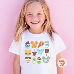 May include: A white short-sleeved t-shirt featuring a colorful graphic print of various food items shaped like Mickey Mouse ears, including a sundae, waffle, pizza slice, coffee cup, burger, candy apple, cotton candy, popsicle, ice cream cone, and donut.