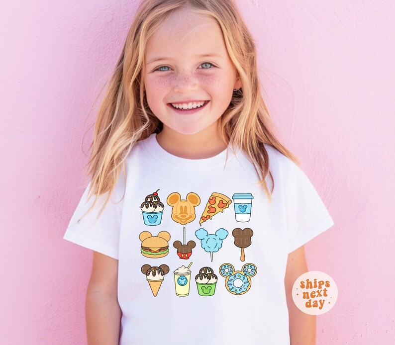 May include: A white short-sleeved t-shirt featuring a colorful graphic print of various food items shaped like Mickey Mouse ears, including a sundae, waffle, pizza slice, coffee cup, burger, candy apple, cotton candy, popsicle, ice cream cone, and donut.