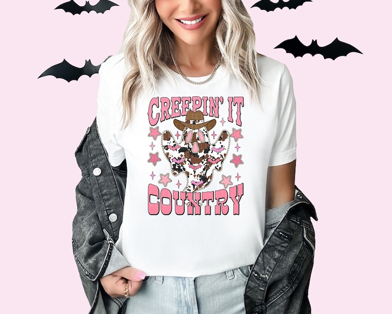 Pode incluir: Camiseta branca com um fantasma rosa e marrom usando um chap&eacute;u de cowboy e o texto "Creepin' It Country".
