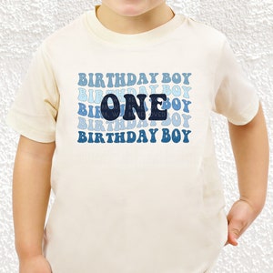 以下が含まれることがあります： 白の半袖Tシャツに、青と白のグラデーションで「Birthday Boy」という文字が複数回繰り返されているデザイン。中央には濃い青色の「One」という数字がデザインされている。