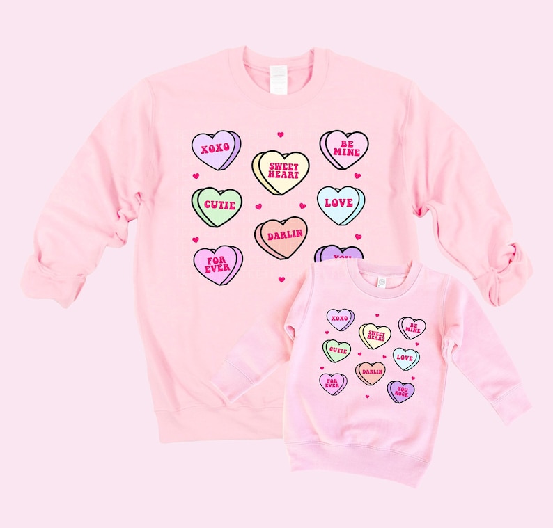 May include: Two pink sweatshirts with a Valentine's Day theme. The larger sweatshirt has a pattern of candy hearts with phrases like "XOXO", "Sweet Heart", "Be Mine", "Gutie", "Love", "Darlin", "For Ever". The smaller sweatshirt has the same pattern but with fewer hearts.