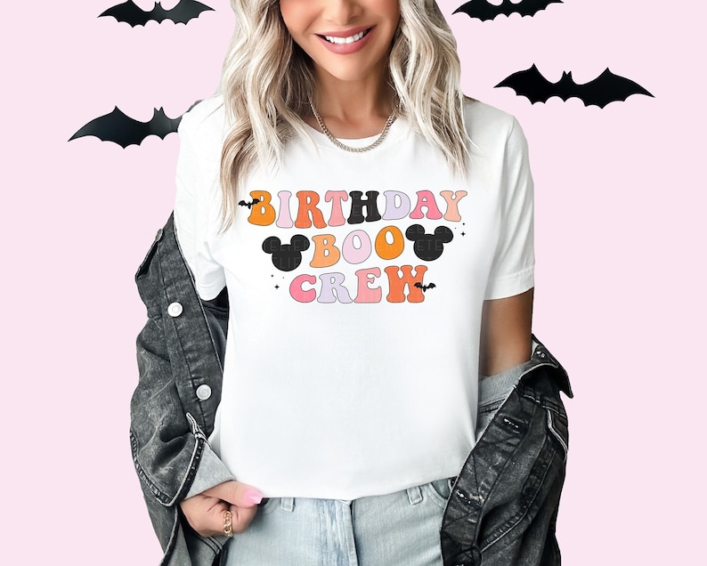 Puede incluir: Camiseta blanca con el texto "Birthday Boo Crew" en letras retro de colores. El texto est&aacute; decorado con murci&eacute;lagos negros y orejas de Mickey Mouse.