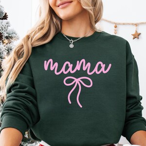 May include: Dark green sweatshirt with the word "mama" in pink script and a pink bow graphic. The sweatshirt has a crew neck and long sleeves. A silver necklace is visible at the neckline.