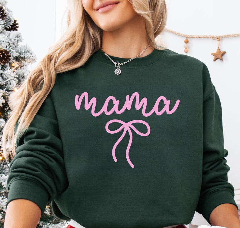 May include: Dark green sweatshirt with the word "mama" in pink script and a pink bow graphic. The sweatshirt has a crew neck and long sleeves. A silver necklace is visible at the neckline.