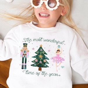 May include: White long-sleeved sweatshirt with a festive Christmas design. The graphic features a nutcracker, a Christmas tree, and a ballerina. The text "The most wonderful time of the year" is displayed on the front.