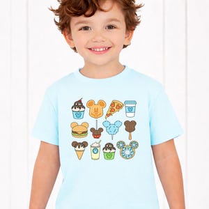 May include: A light blue short-sleeve t-shirt featuring a grid of colorful food illustrations, including a Mickey Mouse-shaped waffle, pizza slice, burger, cotton candy, ice cream cone, donut, and various drinks, all with small Mickey icons.