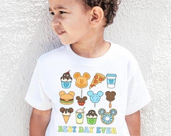 Peut inclure: T-shirt blanc avec des icônes de nourriture de dessin animé colorées, notamment de la crème glacée, des gaufres, des pizzas et une tasse de café. Le t-shirt porte également l'inscription "BEST DAY EVER" en vert et bleu.