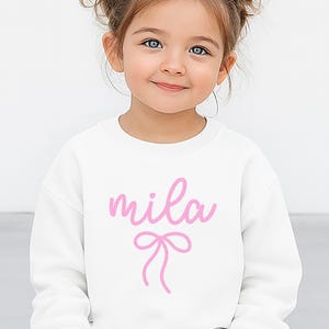 May include: A white sweatshirt with the name "mila" in pink cursive script and a pink bow graphic. The sweatshirt is a crew neck style and appears to be made of a soft material. The design is centered on the front of the garment.