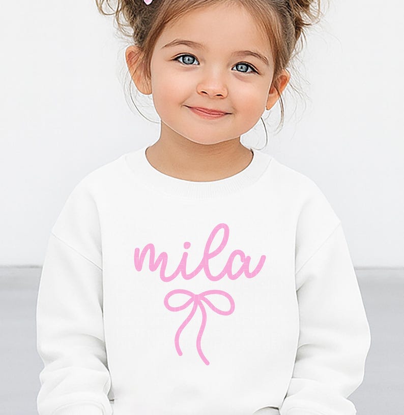 May include: A white sweatshirt with the name "mila" in pink cursive script and a pink bow graphic. The sweatshirt is a crew neck style and appears to be made of a soft material. The design is centered on the front of the garment.
