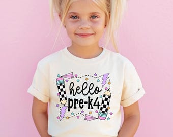 May include: A cream-colored short-sleeve t-shirt featuring a graphic print with the text "hello pre-k4" in black script. The design includes two checkered pencils, pink paper airplanes, purple lightning bolts, and small colorful stars.