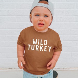 May include: A short-sleeved, rust-colored crew neck t-shirt featuring the white, distressed-style text "WILD TURKEY" printed across the chest. The shirt is paired with a light gray baseball cap worn backwards, completing the casual look.