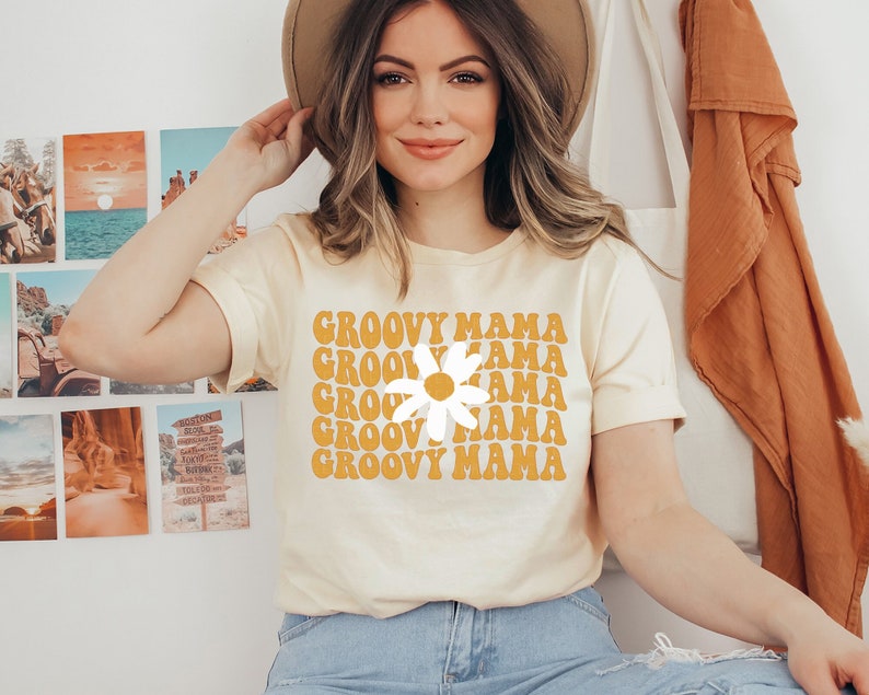 May include: A light yellow t-shirt with the words "GROOVY MAMA" in orange, stacked vertically with a white daisy in the center. The shirt is being worn by a person. The person is wearing a brown hat and blue jeans.