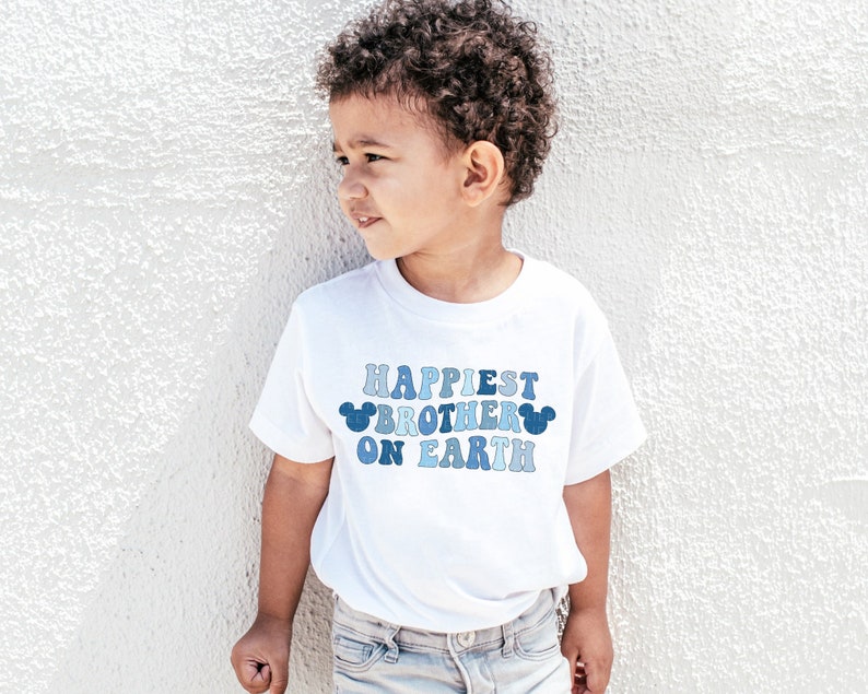 May include: A white t-shirt with a blue and white graphic that says "Happiest Brother on Earth" with Mickey Mouse ears on either side of the text.