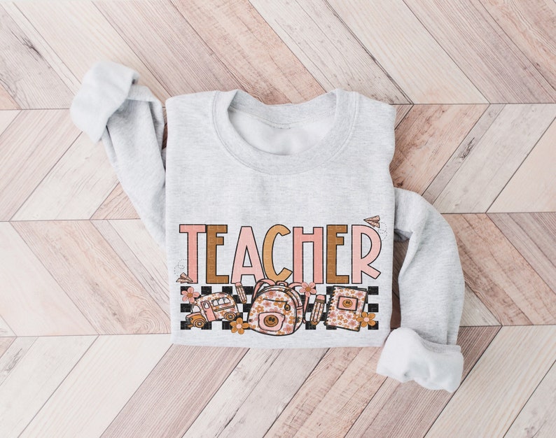 Puede incluir: Una sudadera gris con la palabra "TEACHER" en letras rosas y marrones. El dise&ntilde;o incluye un autob&uacute;s escolar, una mochila y libros con detalles florales.
