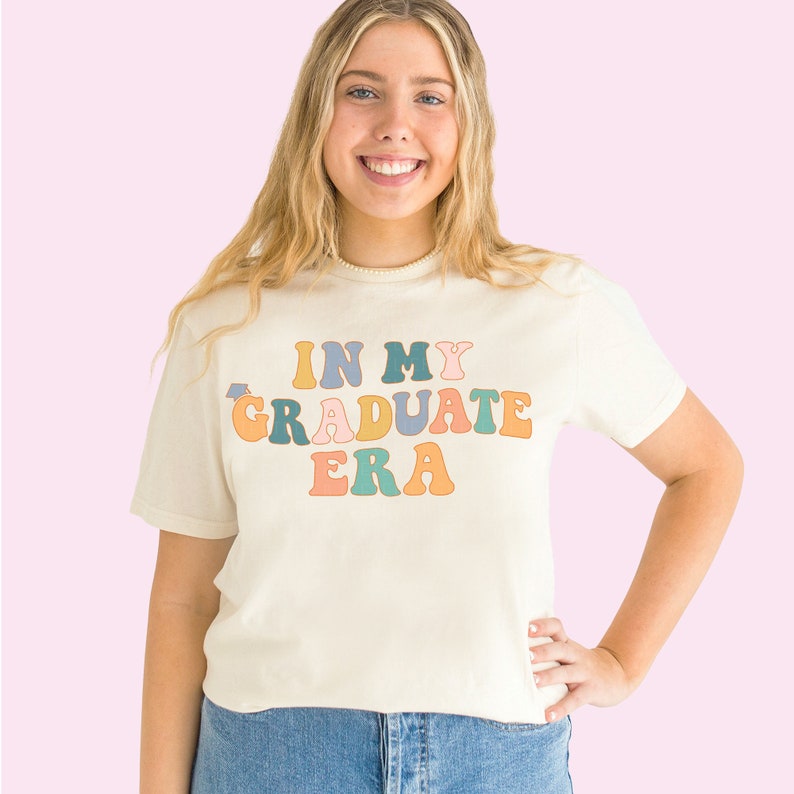 K&ouml;nnte beinhalten: Ein wei&szlig;es T-Shirt mit dem Text "IN MY GRADUATE ERA" in bunten Retro-Lettern.