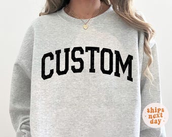 May include: A light gray sweatshirt with the word "CUSTOM" in bold, black, block letters across the chest. The sweatshirt has a crew neck and long sleeves. The image also includes a small, round sticker that says "ships next day."