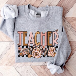 Puede incluir: Sudadera gris con la palabra "TEACHER" en letras rosa y marr&oacute;n. La palabra est&aacute; rodeada de un patr&oacute;n a cuadros e im&aacute;genes de un autob&uacute;s escolar, una mochila y un cuaderno con dise&ntilde;os florales.