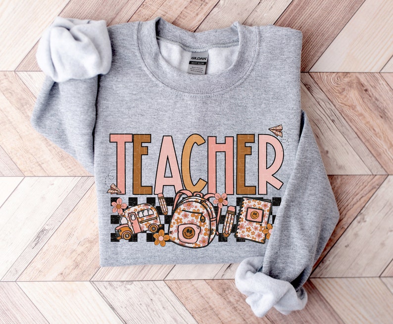 Puede incluir: Sudadera gris con la palabra "TEACHER" en letras rosa y marr&oacute;n. La palabra est&aacute; rodeada de un patr&oacute;n a cuadros e im&aacute;genes de un autob&uacute;s escolar, una mochila y un cuaderno con dise&ntilde;os florales.