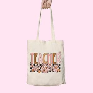 Puede incluir: Una bolsa de tela blanca con la palabra "TEACHER" impresa en letras rosas y marrones. La bolsa presenta un autob&uacute;s escolar, una mochila y libros con motivos florales.