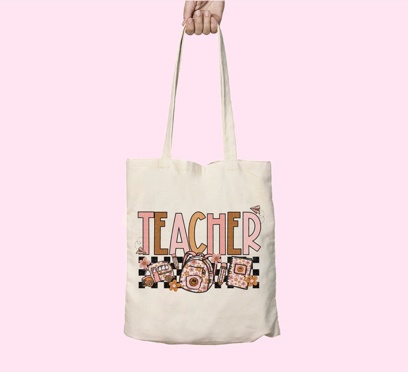 Puede incluir: Una bolsa de tela blanca con la palabra "TEACHER" impresa en letras rosas y marrones. La bolsa presenta un autob&uacute;s escolar, una mochila y libros con motivos florales.