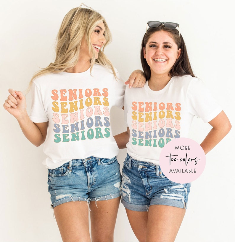 May include: Two women wearing white t-shirts with the word "Seniors" repeated in a retro font in various pastel colors. The text is stacked vertically on the shirts. The women are smiling and looking at the camera. The text "More Tee Colors Available" is in a circle on the bottom right of the image.