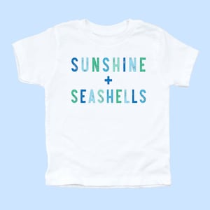 May include: White t-shirt with the text "Sunshine + Seashells" in blue, green, and light blue lettering.