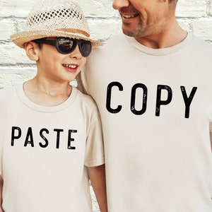 May include: Two people wearing matching white t-shirts with black text. The adult's shirt says "COPY" and the child's shirt says "PASTE".