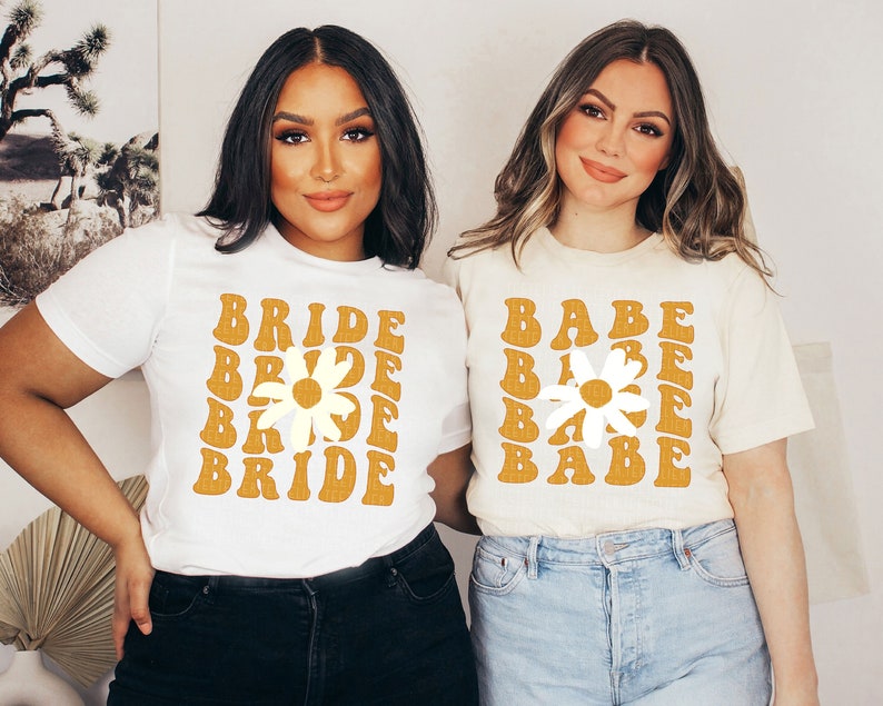 K&ouml;nnte beinhalten: Zwei Personen tragen wei&szlig;e T-Shirts mit einem G&auml;nsebl&uuml;mchen-Motiv und den Worten "Bride" und "Babe" in einer Retro-Schriftart. Die Shirts sind cremefarben mit einem gelben G&auml;nsebl&uuml;mchen-Motiv.