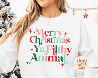 May include: White sweatshirt with the text "Merry Christmas Ya Filthy Animal" in red, green, pink, and teal. The text is decorated with star accents. The sweatshirt is a festive holiday garment.