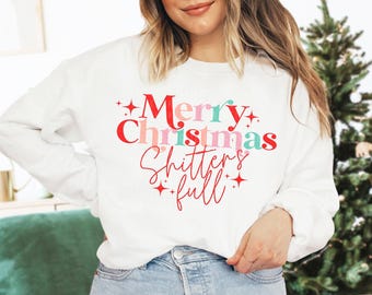 May include: White long-sleeve sweatshirt with the text "Merry Christmas Shitters full" in red, pink, and teal. The text is in a playful font with star accents, perfect for the holiday season.