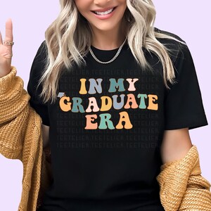 K&ouml;nnte beinhalten: Ein schwarzes T-Shirt mit dem Text "In My Graduate Era" in bunten, Retro-Lettern.