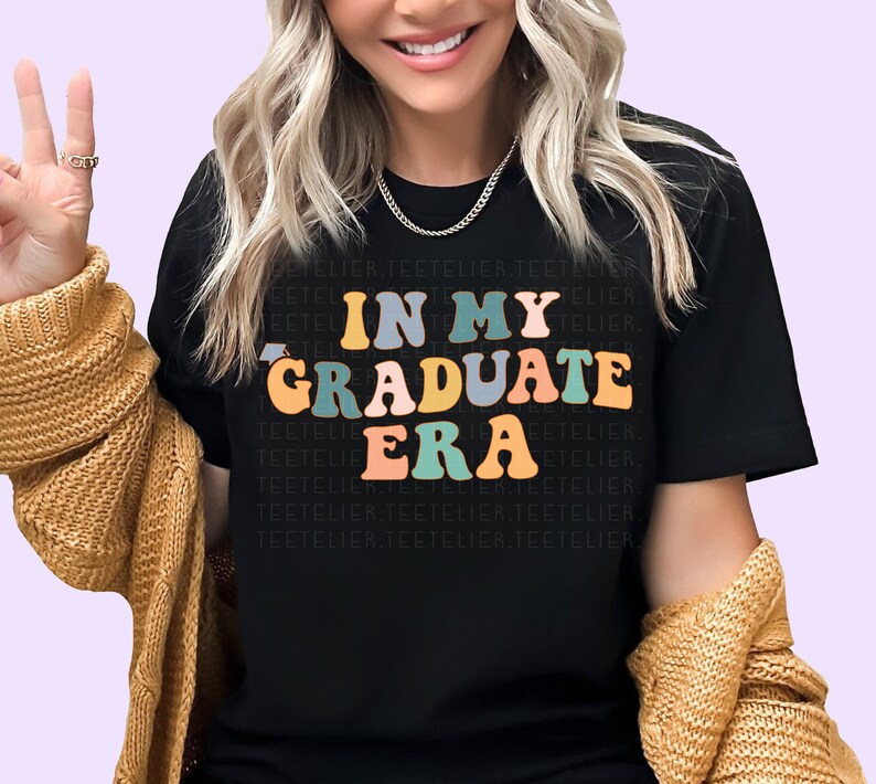 K&ouml;nnte beinhalten: Ein schwarzes T-Shirt mit dem Text "In My Graduate Era" in bunten, Retro-Lettern.